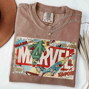Puede incluir: Una camiseta marr&oacute;n Comfort Colors con un dise&ntilde;o de c&oacute;mic de Marvel. El gr&aacute;fico incluye el logotipo de Marvel, vi&ntilde;etas de c&oacute;mic y palabras de acci&oacute;n. Un collar plateado con un colgante descansa sobre la camiseta.