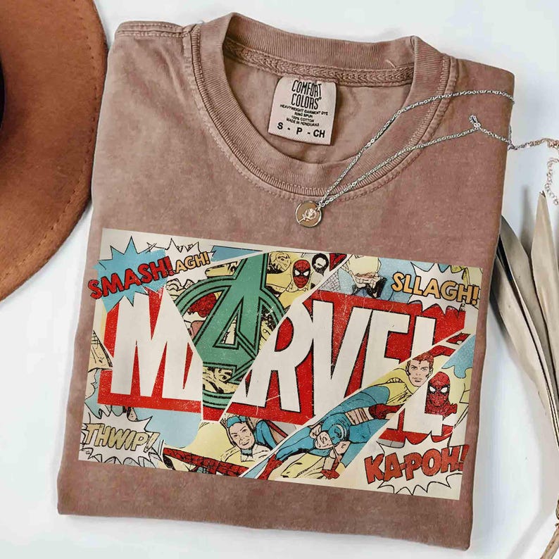 Puede incluir: Una camiseta marr&oacute;n Comfort Colors con un dise&ntilde;o de c&oacute;mic de Marvel. El gr&aacute;fico incluye el logotipo de Marvel, vi&ntilde;etas de c&oacute;mic y palabras de acci&oacute;n. Un collar plateado con un colgante descansa sobre la camiseta.