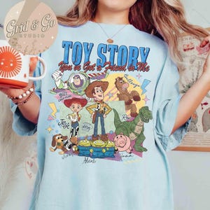 May include: Light blue t-shirt featuring the text "TOY STORY" and "You've Got a Friend in Me". The shirt has a graphic of characters including Buzz Lightyear, Woody, Jessie, Rex, and the Aliens. The shirt is a comfortable, casual wear.