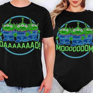 May include: Black t-shirts featuring a cartoon graphic of three green aliens with three eyes, wearing blue and purple outfits, inside a blue circle. One shirt says "DAAAAAAAD!" and the other says "MOOOOOOOM!"