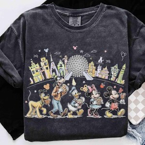 May include: Dark gray t-shirt featuring a colorful graphic of Disney characters, including Mickey Mouse, Minnie Mouse, Goofy, Donald Duck, and Pluto, walking in front of iconic landmarks. The shirt has a vintage, washed look.