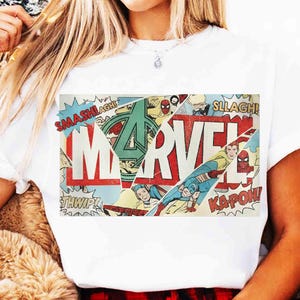 Puede incluir: Camiseta blanca con un dise&ntilde;o gr&aacute;fico de c&oacute;mic de Marvel. El dise&ntilde;o presenta el logotipo de Marvel en rojo y blanco, junto con paneles de c&oacute;mic con personajes como el Capit&aacute;n Am&eacute;rica y Spider-Man, y palabras de acci&oacute;n.