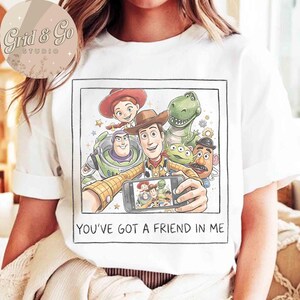 May include: White t-shirt featuring a colorful graphic of characters from the animated movie Toy Story. The graphic shows Woody taking a selfie with Buzz Lightyear, Rex, Jessie, Mr. Potato Head, and the aliens. The text "YOU'VE GOT A FRIEND IN ME" is printed below the graphic.