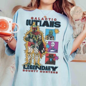 May include: Pale blue t-shirt with the words "GALACTIC OUTLAWS LEGENDARY BOUNTY HUNTERS" and a comic-style illustration of a bounty hunter and three other characters. The design includes yellow lightning bolts.