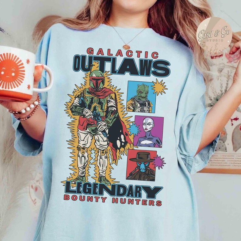 May include: Pale blue t-shirt with the words "GALACTIC OUTLAWS LEGENDARY BOUNTY HUNTERS" and a comic-style illustration of a bounty hunter and three other characters. The design includes yellow lightning bolts.