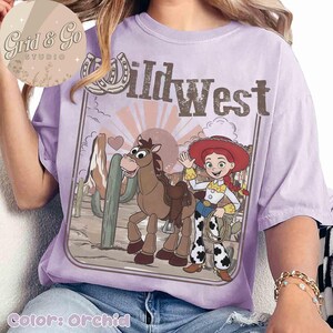 May include: Orchid-coloured t-shirt with a Wild West design. The graphic features a cartoon horse, a cowgirl, cacti, and the text "Wild West." The design is framed by a horseshoe and a heart.