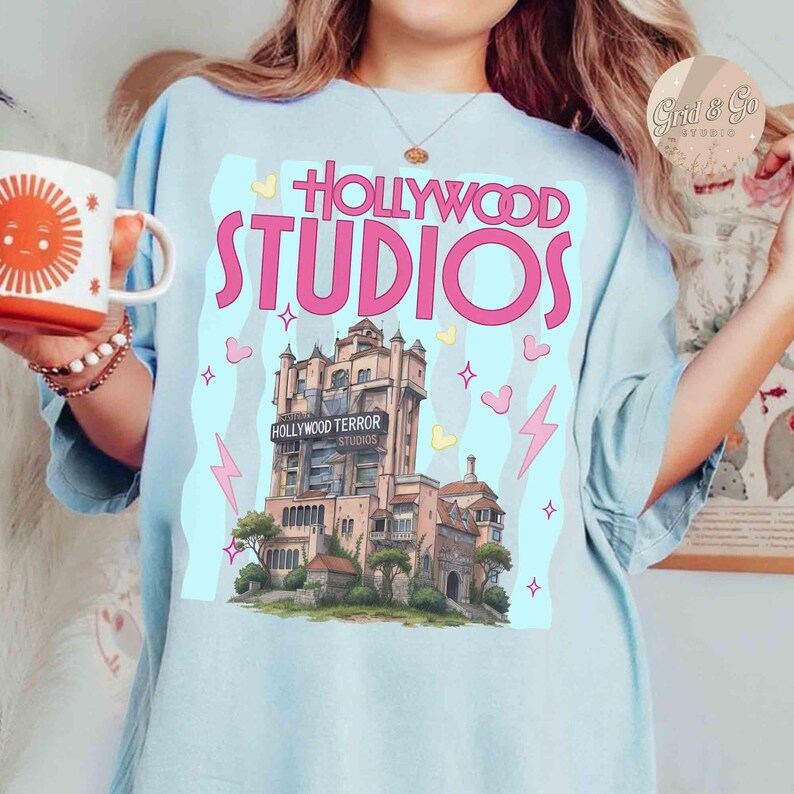 Pu&ograve; includere: T-shirt azzurro con grafica rosa del logo Hollywood Studios e dell'Hollywood Tower Hotel. Il design include cuori, stelle e fulmini rosa. La maglietta ha una vestibilit&agrave; ampia.