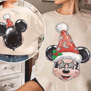 Könnte beinhalten: Ein beigefarbenes Sweatshirt mit Rundhalsausschnitt und einer Grafik eines Cartoon-Mauskopfes, der eine rote Weihnachtsmannmütze mit Mond, Sternen, Stechpalmenblättern und sternförmiger Sonnenbrille trägt. Die Rückansicht zeigt einen schwarzen Luftballon.