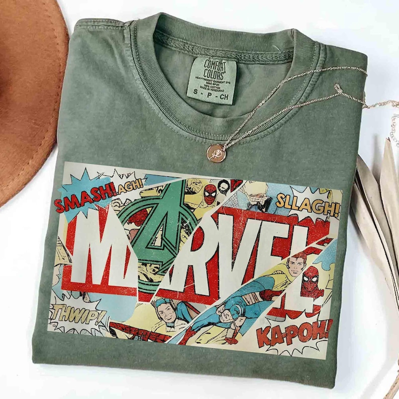 Puede incluir: Camiseta verde oliva con un dise&ntilde;o gr&aacute;fico de c&oacute;mic de Marvel. El gr&aacute;fico presenta el logotipo de Marvel, vi&ntilde;etas de c&oacute;mic y palabras de acci&oacute;n como "SMASH!" y "KAPOW!". Un collar plateado con un peque&ntilde;o colgante descansa sobre la camiseta.