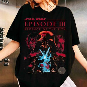 May include: A black t-shirt featuring a graphic print from Star Wars Episode III Revenge of the Sith. The design includes red-toned portraits of Anakin Skywalker and Obi-Wan Kenobi, Darth Vader's helmet, and two figures dueling with blue lightsabers.