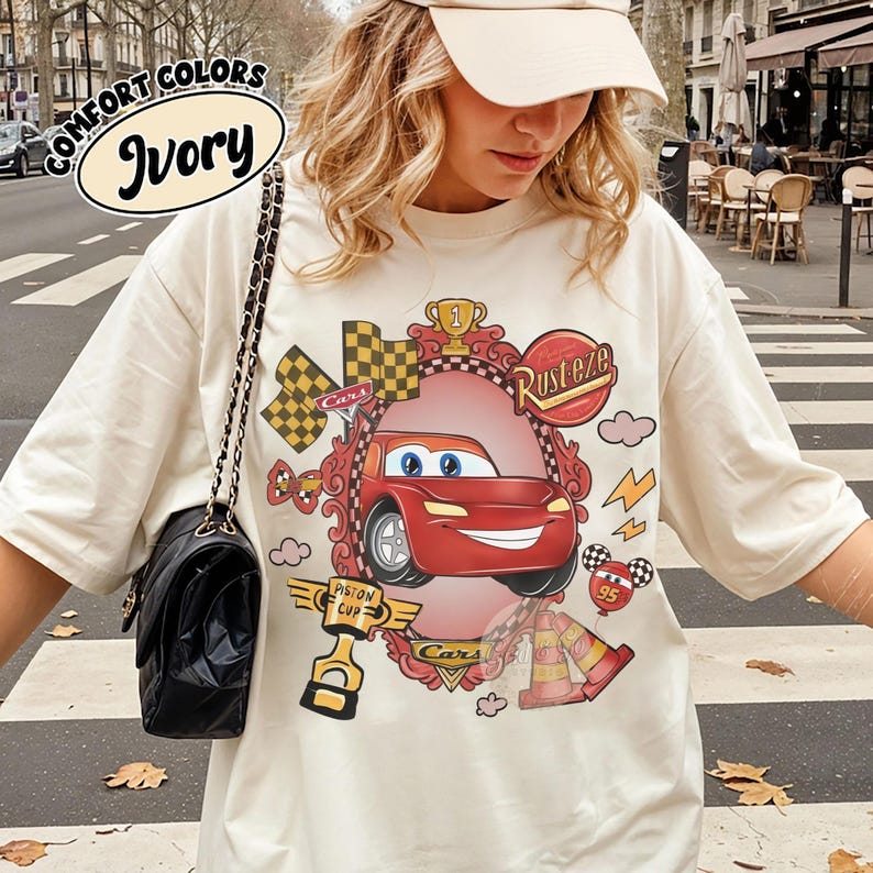 Puede incluir: Camiseta de color marfil con una imagen de dibujos animados de un coche de carreras rojo, el texto "Rusteze" y "Cars". El dise&ntilde;o incluye banderas a cuadros, un trofeo y rayos. La camiseta tambi&eacute;n tiene el texto "Comfort Colors" e "Ivory".