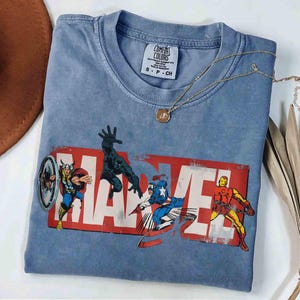 Puede incluir: Una camiseta azul Comfort Colors con el logo de Marvel y los personajes de c&oacute;mic Thor, Black Panther, Capit&aacute;n Am&eacute;rica e Iron Man. Un collar dorado con un colgante est&aacute; sobre la camiseta.