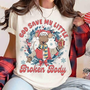 May include: Cream-colored t-shirt featuring a cartoon mouse character in a Santa hat and red jacket, holding a gift. The shirt has the text "GOD SAVE MY LITTLE BROKEN BODY" in a festive design with a wreath and snowflakes.