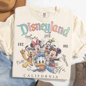 Puede incluir: Camiseta color crema con la palabra "Disneyland" en colores arco&iacute;ris, con personajes de dibujos animados como Mickey Mouse, Minnie Mouse y Goofy. La camiseta incluye el texto "EST. 1955" y "CALIFORNIA".