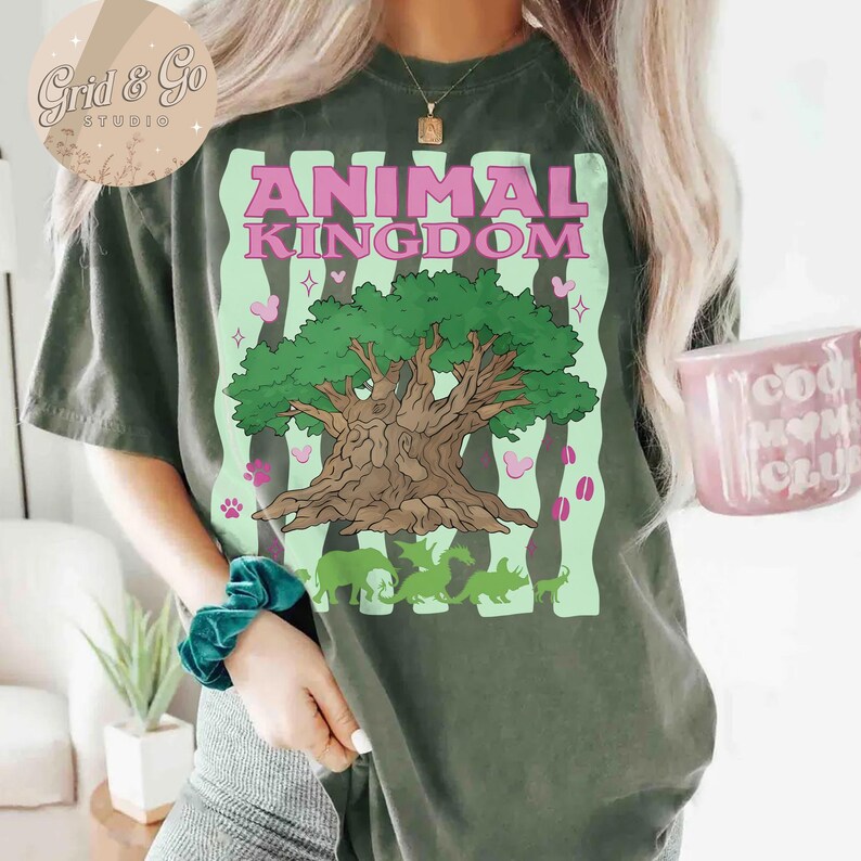 Pu&ograve; includere: T-shirt verde scuro con la scritta "ANIMAL KINGDOM" in rosa, con una grafica ad albero e sagome di animali verdi. La maglietta ha un aspetto vintage.