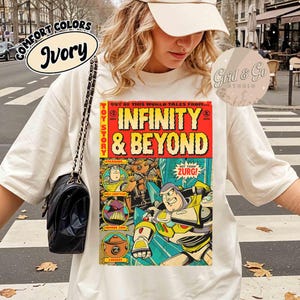 May include: Ivory-colored t-shirt featuring a comic book-style graphic with the text "INFINITY & BEYOND" and characters from Toy Story. The shirt also has the text "COMFORT COLORS" and "Grid & Go STUDIO".