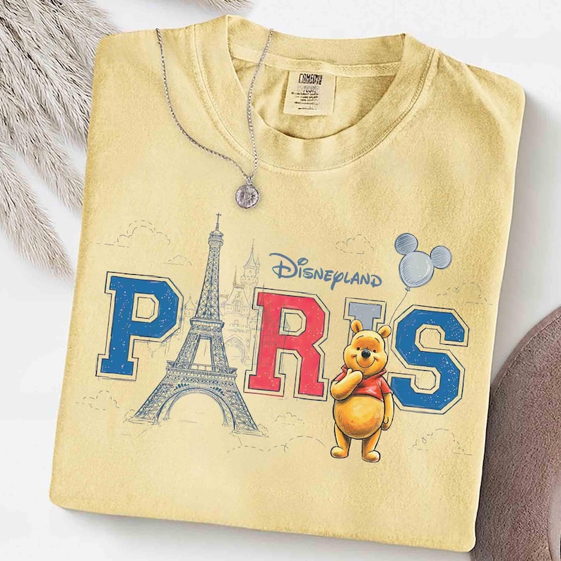 K&ouml;nnte beinhalten: Hellgelbes T-Shirt mit dem Schriftzug "Disneyland Paris" in Blau und Rot, mit dem Eiffelturm und Winnie Puuh. Eine silberne Halskette mit Anh&auml;nger liegt auf dem Shirt.