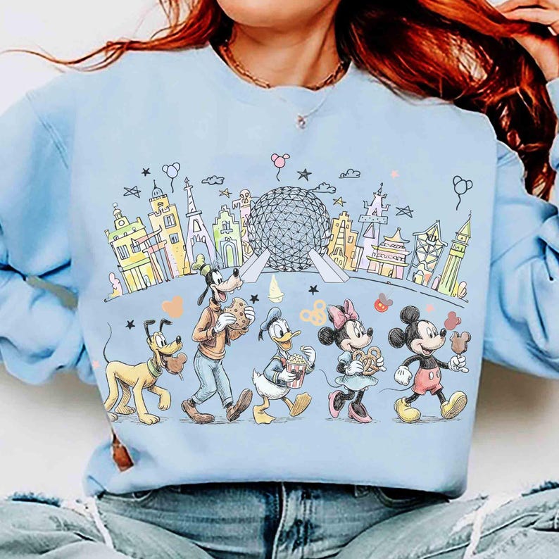 May include: Light blue sweatshirt featuring a colorful illustration of Disney characters, including Mickey Mouse, Minnie Mouse, Donald Duck, Goofy, and Pluto, walking in front of a stylized cityscape with a large sphere. The design includes hearts and stars.