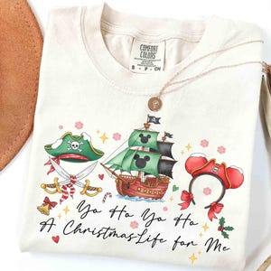 Könnte beinhalten: Cremefarbenes T-Shirt mit einem Piraten-Weihnachtsdesign. Die Grafik zeigt ein Piratenschiff, einen Piratenhut und ein Stirnband mit Mickey Mouse Ohren. Der Text lautet "Yo Ho Yo Ho A Christmas Life for Me."