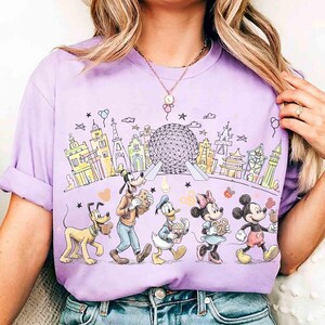 May include: A lilac-colored t-shirt featuring a colorful illustration of Disney characters walking in front of a cityscape. Pluto, Goofy, Donald Duck, Minnie Mouse, and Mickey Mouse are depicted with food items. The background includes a large sphere and various buildings.