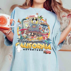 May include: Light blue t-shirt featuring a colorful graphic of Disney California Adventure. The design includes Mickey Mouse, a Ferris wheel, and various other Disney characters. The text "Disney California Adventure" is prominently displayed.