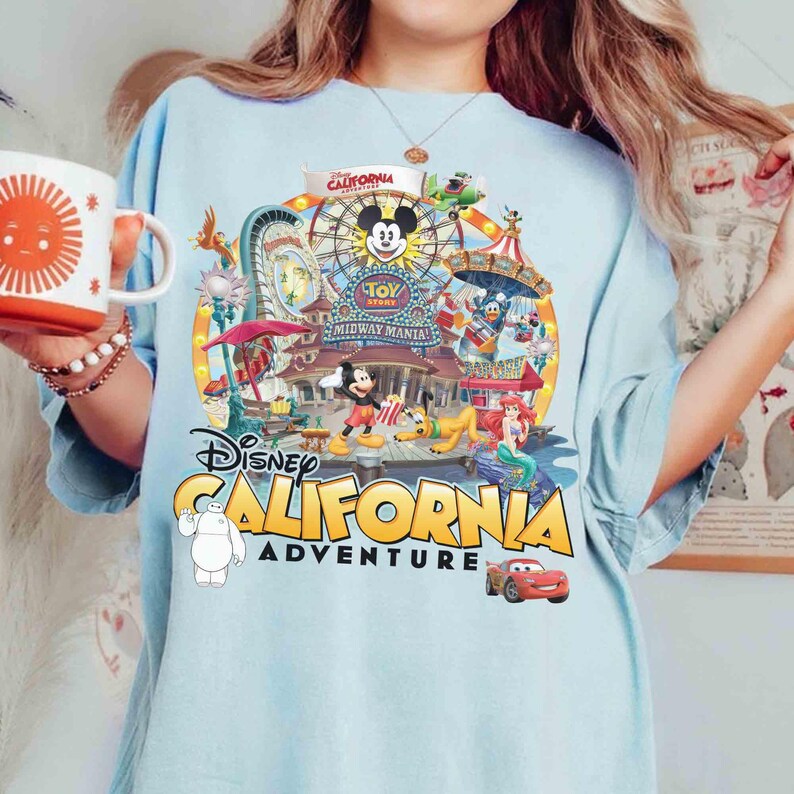 May include: Light blue t-shirt featuring a colorful graphic of Disney California Adventure. The design includes Mickey Mouse, a Ferris wheel, and various other Disney characters. The text "Disney California Adventure" is prominently displayed.