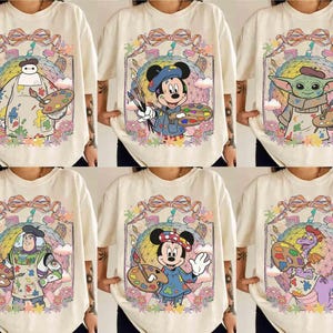 Puede incluir: Camisetas color crema con varios personajes de Disney como artistas. Cada camiseta muestra un personaje diferente, incluyendo Baymax, Mickey Mouse, Grogu, Buzz Lightyear, Minnie Mouse y Figment, sosteniendo paletas y pinceles dentro de un marco colorido.