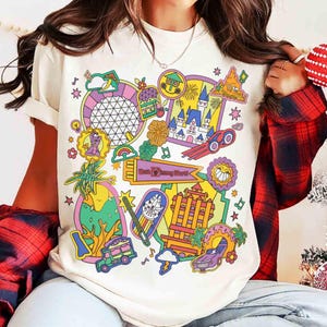 May include: Cream-colored t-shirt featuring a colorful graphic design with various illustrations, including a castle, a spaceship, and the text "Walt Disney World". The design incorporates elements like rainbows, stars, and a sun, creating a vibrant and playful aesthetic.