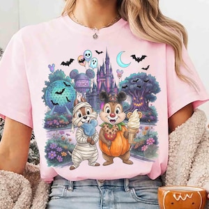 May include: A light pink t-shirt featuring a graphic print of two chipmunks in Halloween costumes, one as a mummy and one in a pumpkin suit. The background shows a theme park castle, a geodesic sphere, a spooky tree, bats, and balloons.