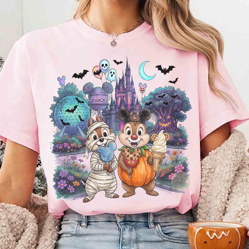 May include: A light pink t-shirt featuring a graphic print of two chipmunks in Halloween costumes, one as a mummy and one in a pumpkin suit. The background shows a theme park castle, a geodesic sphere, a spooky tree, bats, and balloons.