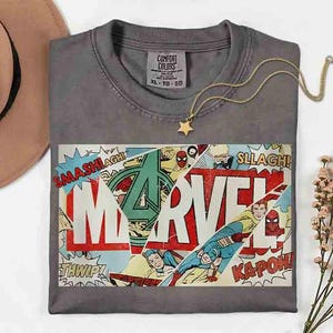 Puede incluir: Camiseta gris con un dise&ntilde;o de c&oacute;mic de Marvel, con el logotipo de Marvel y palabras de c&oacute;mic. Un collar con colgante de estrella dorada descansa sobre la camiseta. Talla XL-TG-EG.
