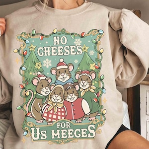 May include: Beige sweatshirt with a festive graphic featuring five cartoon mice wearing holiday attire. The design includes the text "NO CHEESES FOR US MEEGES" surrounded by Christmas lights, trees, and snowflakes. A cozy and whimsical holiday-themed garment.