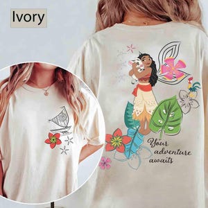 May include: Ivory-colored t-shirt featuring a cartoon illustration of a sailboat and flowers on the front. The back of the shirt displays a character holding a pig, surrounded by flowers and the text "Your adventure awaits."