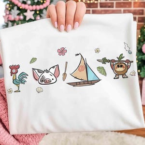 Puede incluir: Una camiseta blanca con un estampado gr&aacute;fico colorido de un gallo de dibujos animados, un lech&oacute;n, un remo de madera, un velero con una vela en espiral y un peque&ntilde;o personaje de coco sosteniendo una lanza, rodeado de flores, hojas y conchas.