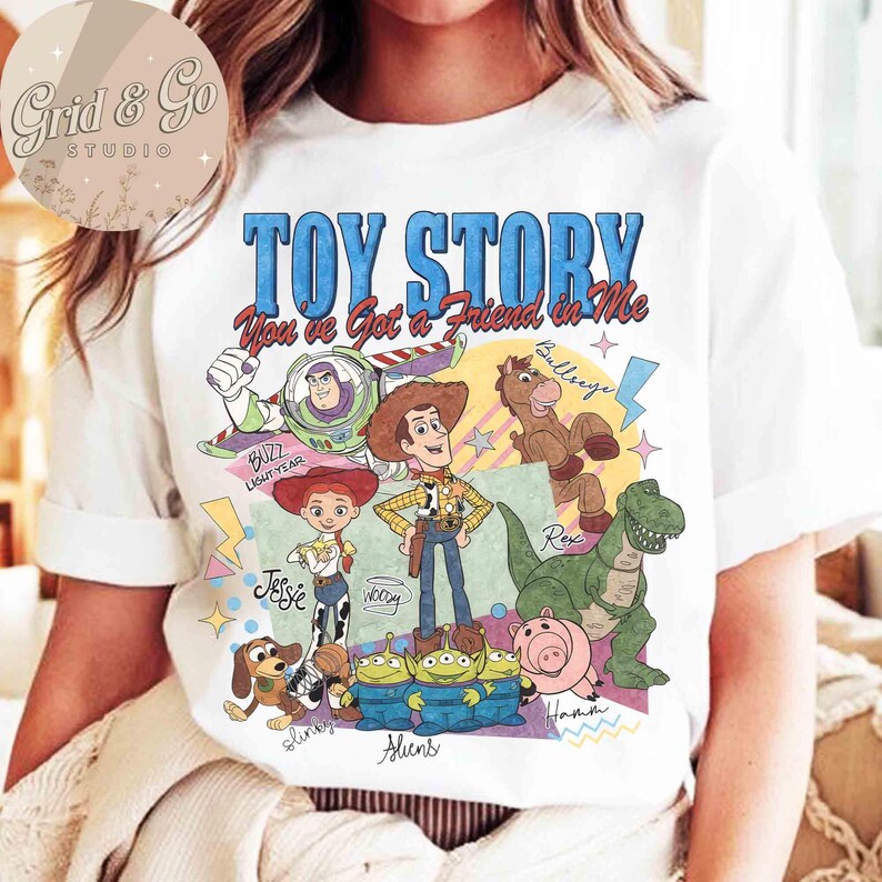 May include: White t-shirt featuring a colorful graphic of characters from the animated film "Toy Story." The design includes Buzz Lightyear, Woody, Jessie, Rex, and other characters, with the text "TOY STORY You've Got a Friend in Me."