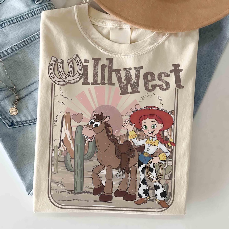 May include: Cream-coloured t-shirt with a Wild West design. The graphic features a cartoon horse, a cowgirl, and the text "Wild West" with horseshoe accents. The shirt is folded on a light blue denim surface.