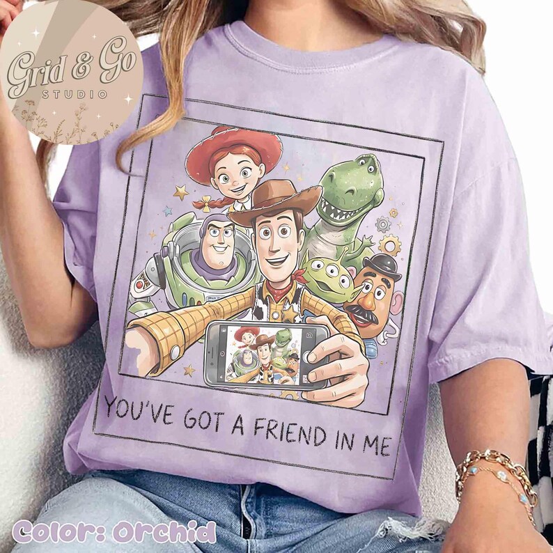 May include: Orchid-colored t-shirt featuring a graphic of Toy Story characters taking a selfie. The image includes Woody, Buzz Lightyear, Rex, Jessie, Mr. Potato Head, and the aliens. The text "YOU'VE GOT A FRIEND IN ME" is printed below the image.