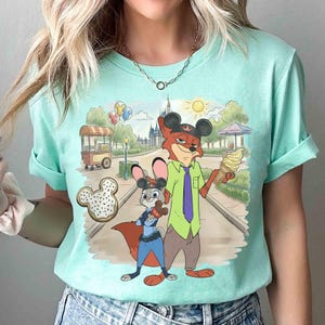 May include: Mint green t-shirt featuring a cartoon scene with a fox and a bunny. The fox wears a hat with mouse ears and holds an ice cream cone. The bunny holds a chocolate treat. A castle and food cart are in the background.