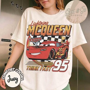 Pu&ograve; includere: T-shirt color crema con grafica di Saetta McQueen del film Cars. Il design include il testo "Lightning McQueen", "Cars", "Think Fast" e il numero "95". La maglietta ha anche il testo "Comfort Colors" e "Juoy".