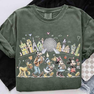May include: Green crew neck sweatshirt featuring a colorful cartoon illustration of Disney characters walking in front of a cityscape. Pluto, Goofy, Donald Duck, Minnie Mouse, and Mickey Mouse are depicted with snacks.