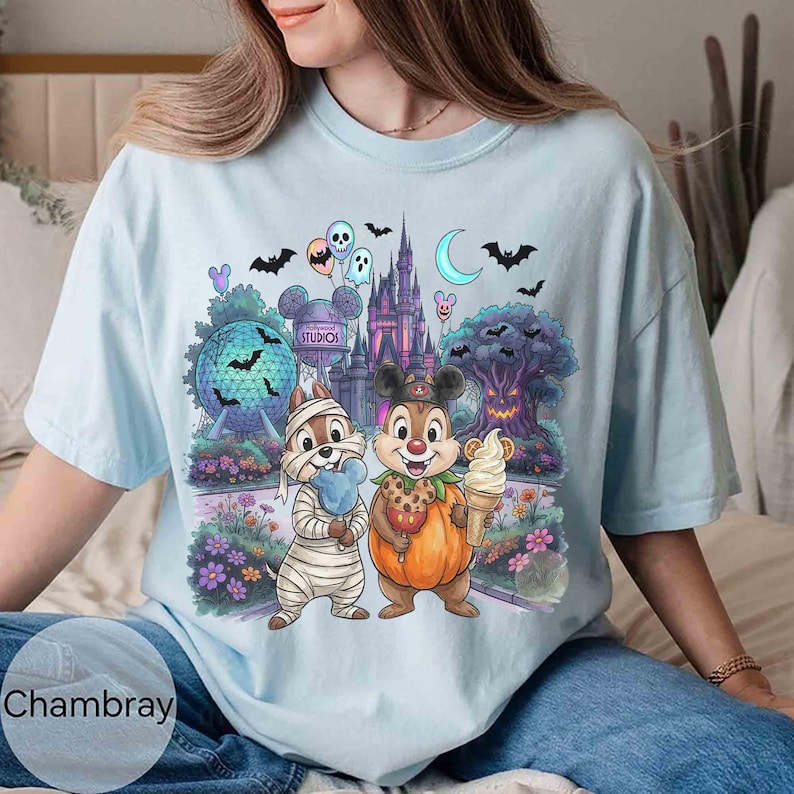 May include: A light blue t-shirt featuring a graphic print of two chipmunks in Halloween costumes, one as a mummy and one as a pumpkin, standing before a theme park castle, a geodesic sphere, and a spooky tree with bats and balloons.