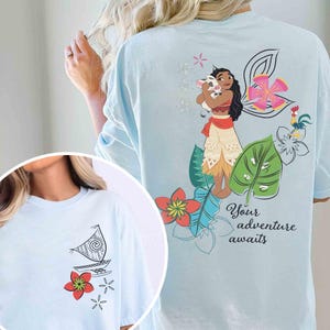 May include: Light blue t-shirt featuring a cartoon illustration of a Polynesian woman holding a pig, surrounded by flowers and the text "Your adventure awaits." A smaller image shows a boat and flowers.