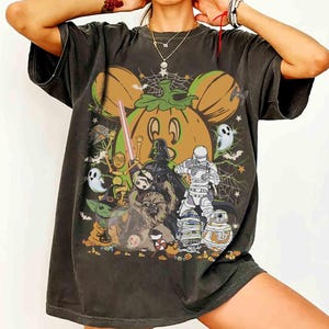 May include: A charcoal gray short-sleeve t-shirt featuring a large graphic print of a pumpkin shaped like mouse ears, surrounded by various science fiction characters, ghosts, spiderwebs, and candy corn in orange, green, white, and black colors.