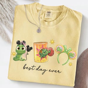 Op de afbeelding: Lichtgele t-shirt met een afbeelding van een groene kameleon, een lantaarn en een hoofdband met een kameleonontwerp. De tekst "best day ever" is in cursief geschreven. Een zilveren ketting met een hanger ligt op het shirt.