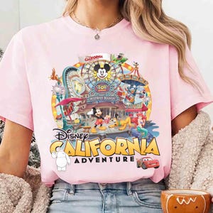 May include: Pink t-shirt featuring a colorful graphic of Disney California Adventure. The design includes Mickey Mouse, a Ferris wheel, and other theme park elements. The words "Disney California Adventure" are prominently displayed.