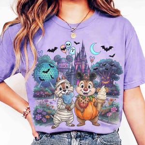 May include: A light purple t-shirt featuring a graphic print of two chipmunks in Halloween costumes, one as a mummy and one as a pumpkin, standing before a theme park castle, a geodesic sphere, and a spooky tree with bats, balloons, and text.