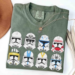 May include: An olive green t-shirt with a graphic of nine stormtrooper helmets. The t-shirt has a crew neck and short sleeves. A gold necklace with a pendant is draped over the shirt.