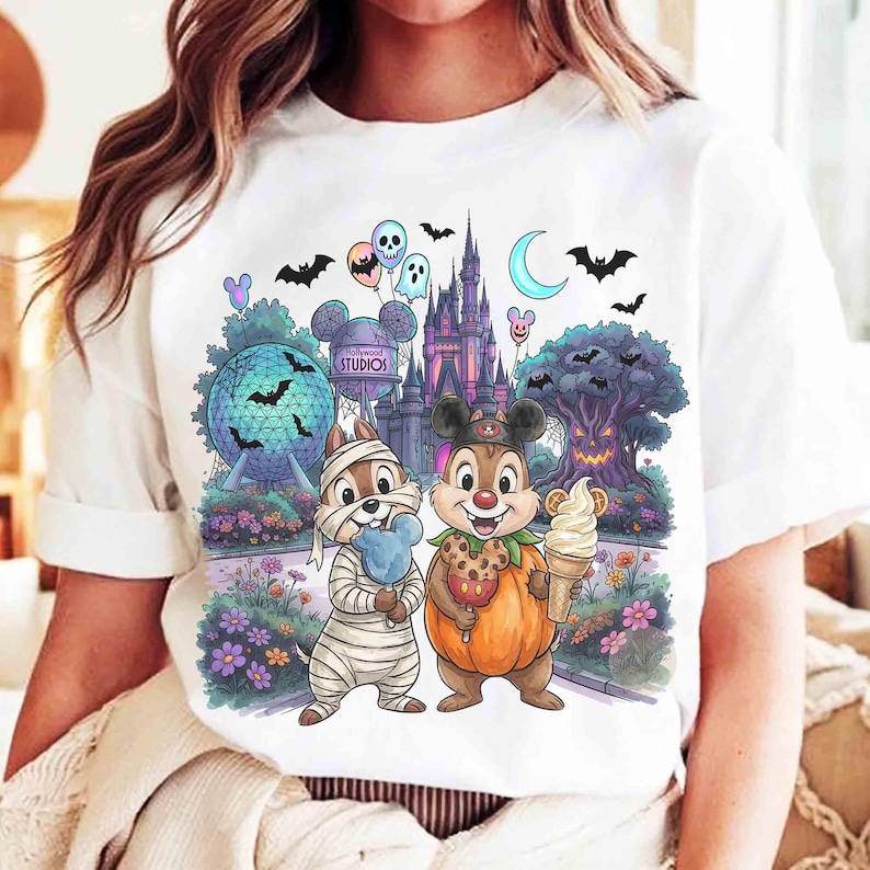 May include: A white t-shirt featuring a colorful graphic of two chipmunks in Halloween costumes, one as a mummy and one in a pumpkin suit, standing before a theme park castle, a geodesic sphere, and a spooky tree with bats and balloons.