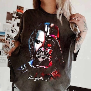 Op de afbeelding: Donkergrijs t-shirt met een gesplitste afbeelding van een persoon en de helm van Darth Vader. Het ontwerp gebruikt rode, blauwe en witte accenten, met een handtekening onderaan. Het shirt heeft een vintage look.