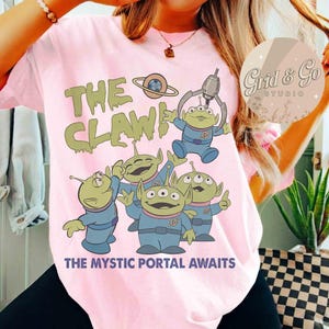 May include: Pink t-shirt featuring a graphic of green aliens in blue suits with the text "THE CLAW!" and "THE MYSTIC PORTAL AWAITS". The aliens are from a popular animated movie.